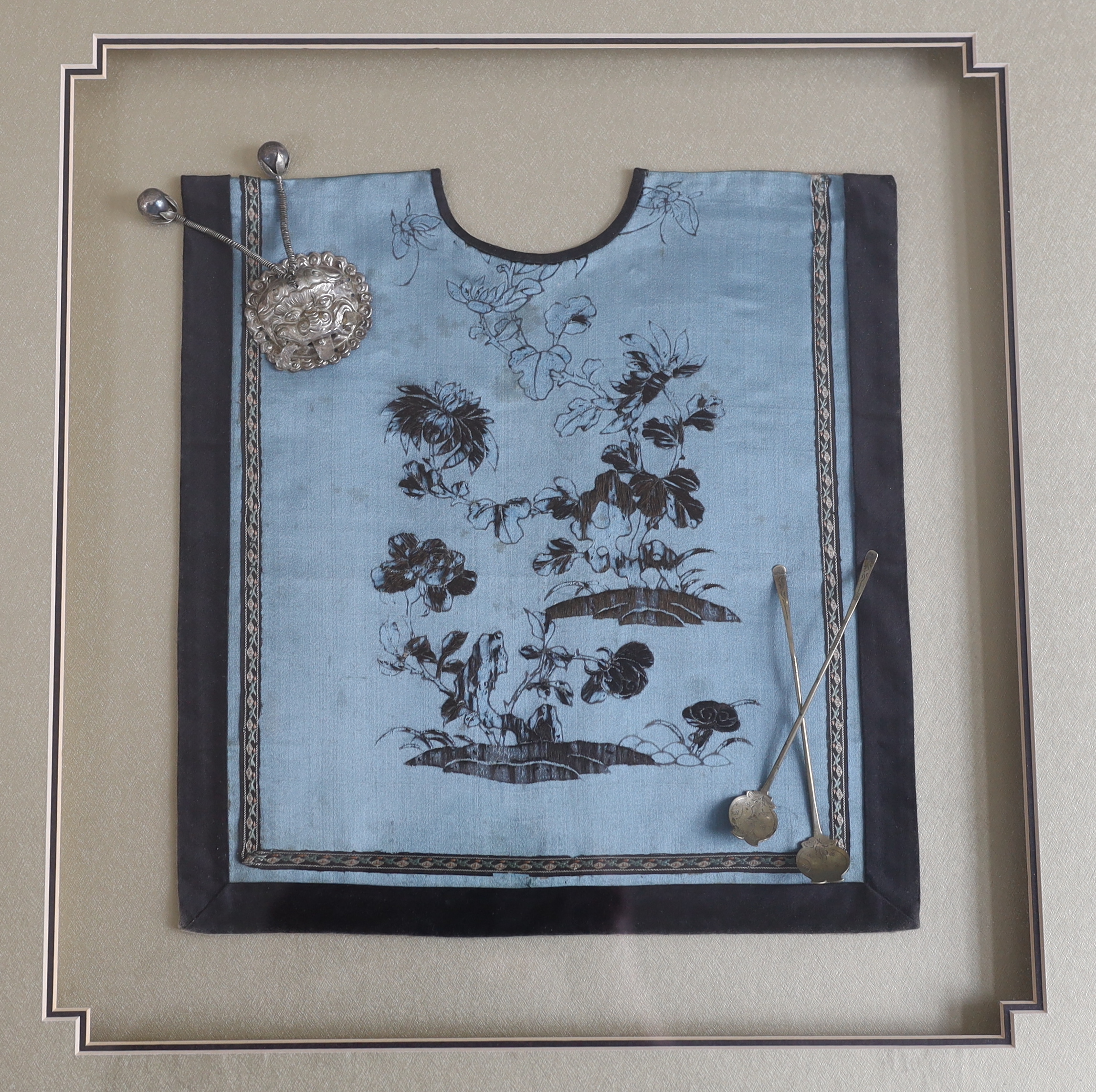 A late 19th century Chinese silk and blue floral embroidered baby’s bib, mounted with two white metal spoons and a rattle, together with a similar bib and spoons plus a decorative white metal mask mount, blue embroidery